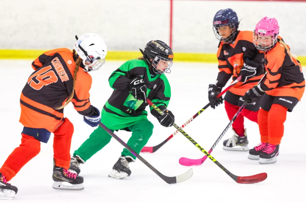Young girls in orange and green jerseys chasing the puck during a game at the 2025 SGHL Mixer.